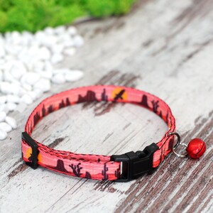 May include: A pink cat collar with a sunset and cactus print, a black buckle, and a red bell.