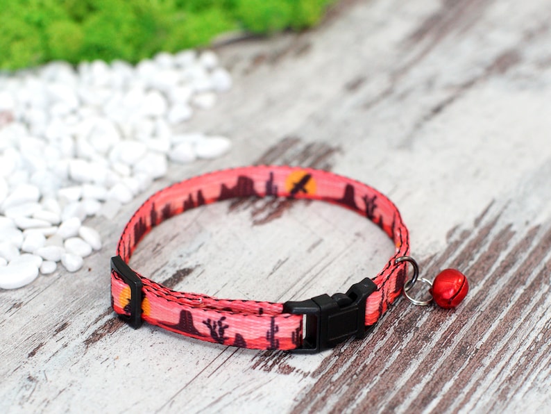 May include: A pink cat collar with a sunset and cactus print, a black buckle, and a red bell.