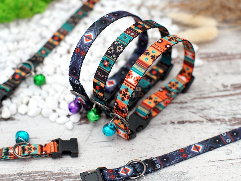 May include: Three colourful cat collars with geometric patterns and bell charms. The collars are made of a soft, woven fabric and feature a black plastic buckle.