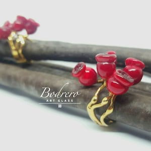 Puede incluir: Dos anillos dorados con diseños de flores de cristal rojo. Los anillos se muestran sobre una superficie texturizada de color marrón oscuro. Los anillos tienen un diseño artístico único, con el texto "Bodrero ART GLASS" en la esquina inferior derecha.