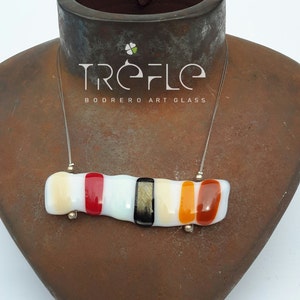 Könnte beinhalten: Eine weiße, orange, rote und schwarze Glaskette mit einer silbernen Kette. Die Kette liegt auf einem braunen Präsentationsständer. Der Text "Treflo Bodrero Art Glass" befindet sich auf dem Ständer.