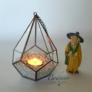 Könnte beinhalten: Ein geometrisches Glas-Terrarium mit einer Kette, die von oben hängt. Das Terrarium ist mit weißen Steinen gefüllt und eine kleine Kerze brennt darin. Eine kleine Keramikfigur eines Mannes in einer gelben Robe und einem schwarzen Hut steht rechts neben dem Terrarium. Der Text "Bodrero Art Glass" ist unten im Bild sichtbar.
