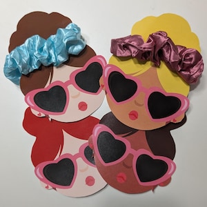 Puede incluir: Recortes de papel de rostros estilizados con diferentes tonos de piel, cada uno con gafas de sol rosas en forma de corazón. Las gomas para el pelo azules y malvas están encima de los recortes. Los rostros tienen labios rojos y están dispuestos en grupo.