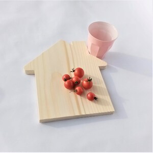 Może przedstawiać: Drewniana deska do krojenia w kształcie domu z jasnym usłojeniem. Kilka czerwonych pomidorków koktajlowych jest rozrzuconych na desce. Różowy, fasetowany kubek jest umieszczony obok deski na białej powierzchni.