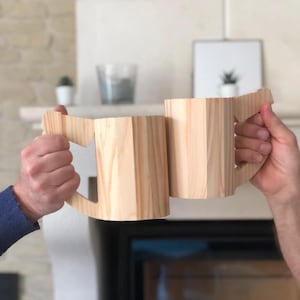 Könnte beinhalten: Zwei Holzbecher mit Henkeln aus hellem braunem Holz werden zum Anstoßen hochgehalten. Die Becher sind scharf, während der Hintergrund unscharf ist.