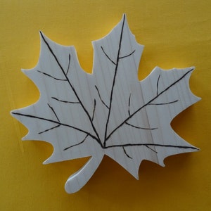 May include: A wooden maple leaf cutout with a black line drawing of veins. The leaf is painted white and is on a yellow background.