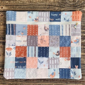 May include: A patchwork zippered pouch with a variety of fabric squares in shades of blue, peach, and gray. The squares feature floral, bird, and text designs. The zipper is white, and the pull has a charm.