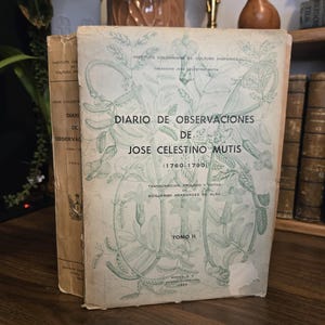 May include: Two vintage books are displayed, one partially obscured. The front book, titled "DIARIO DE OBSERVACIONES DE JOSE CELESTINO MUTIS," features a light green floral design. The text is in Spanish. The book is from the Instituto Colombiano de Cultura Hispánica.