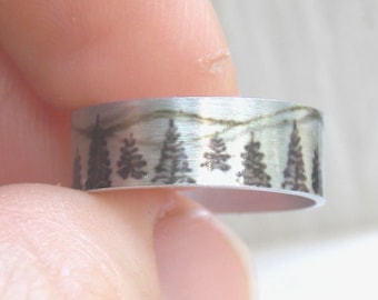 Puede incluir: Un anillo de banda de plata con un acabado cepillado y un diseño grabado de una cordillera con árboles.