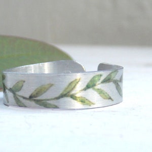 May include: A silver ring with a green leafy design. The ring is made of metal and has a simple, elegant design.
