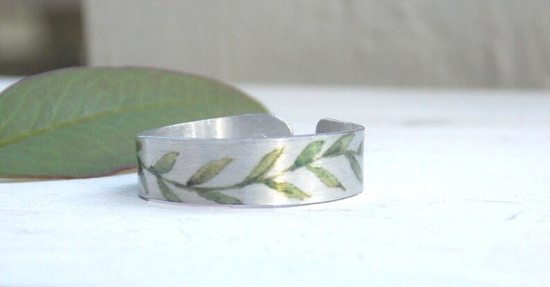 May include: A silver ring with a green leafy design. The ring is made of metal and has a simple, elegant design.