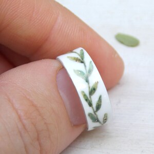 May include: A white ring with a green leafy design. The ring is decorated with a band of green leaves and gold glitter.