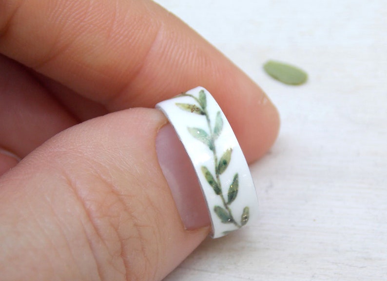 May include: A white ring with a green leafy design. The ring is decorated with a band of green leaves and gold glitter.