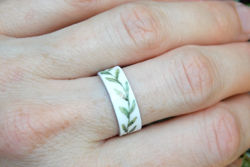 May include: A white ring with a green leafy design. The leaves are painted in shades of green and have a watercolor effect.