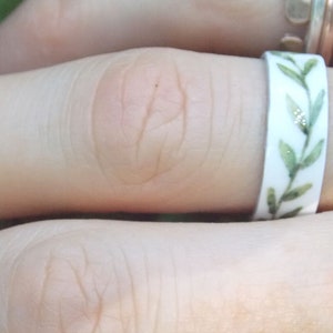 May include: A white ring with a green leafy design. The ring is on a person's finger.