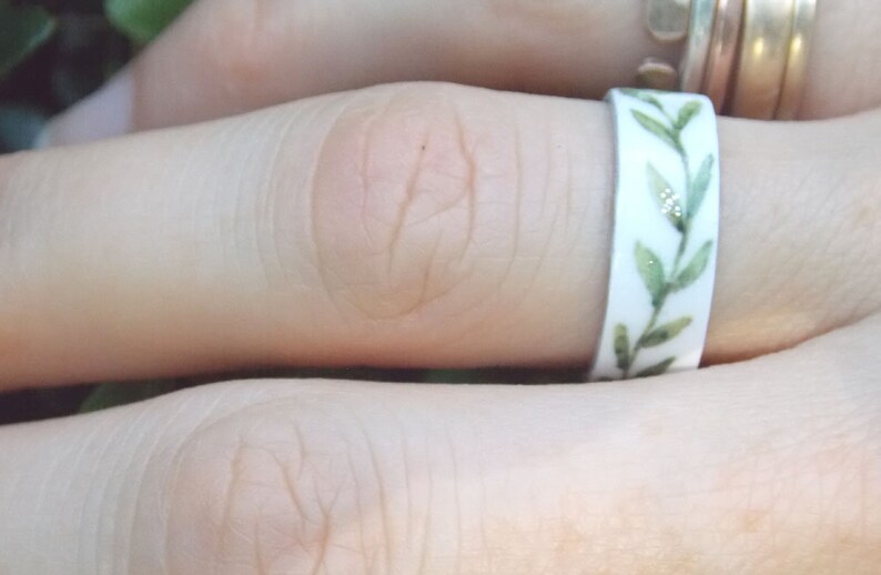 May include: A white ring with a green leafy design. The ring is on a person's finger.