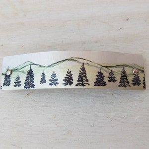 May include: A silver metal bar hair clip with a painted design of a mountain range and a row of pine trees in black and green.