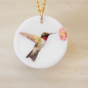 May include: A white ceramic pendant necklace with a gold chain features a hummingbird with iridescent wings and a red throat, drinking nectar from a pink flower.