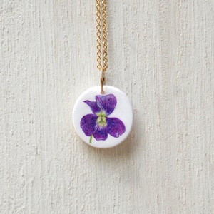 May include: A gold chain necklace with a round pendant featuring a purple flower with green leaves on a white background.