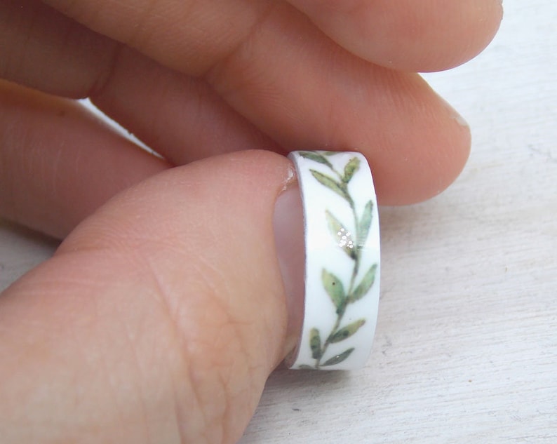 May include: A white ring with a green leafy design. The ring is made of washi tape and has a glittery finish.