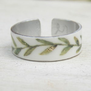 May include: A white ring with a green leafy design and gold accents. The ring is made of metal and has a simple, elegant design.