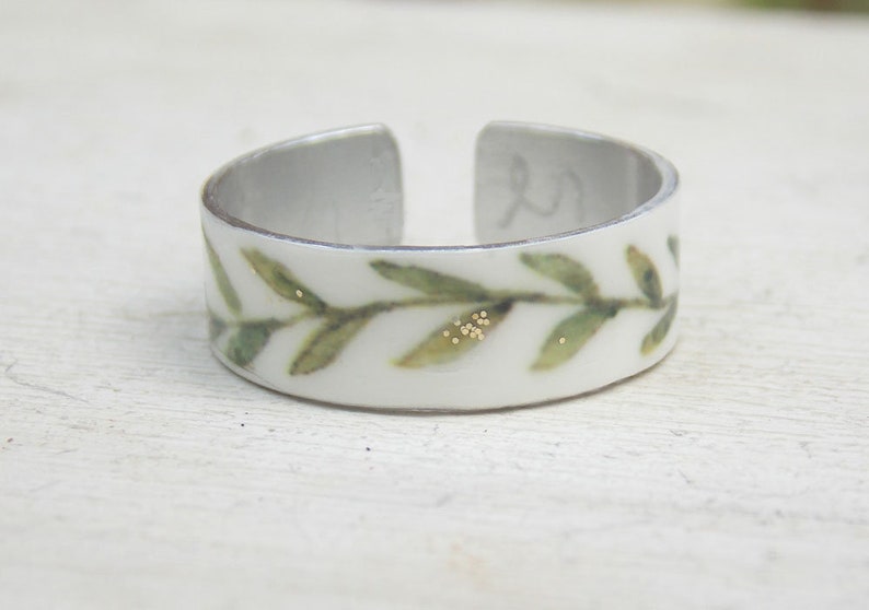 May include: A white ring with a green leafy design and gold accents. The ring is made of metal and has a simple, elegant design.