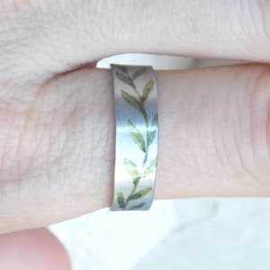May include: A silver ring with a delicate green floral design. The ring is worn on a finger.