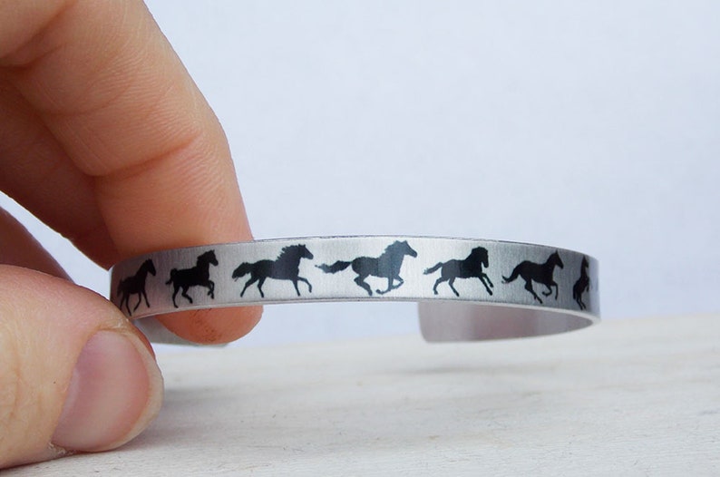 May include: A silver metal bracelet with a repeating pattern of black running horse silhouettes.