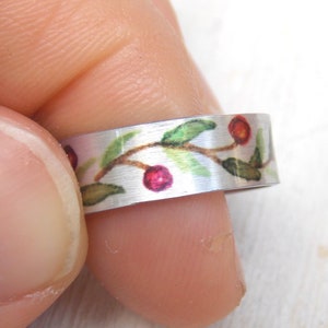 May include: A silver ring with a floral design featuring green leaves and red berries. The ring is worn on a finger.