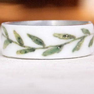 May include: A white ceramic ring with a green leafy design. The leaves have a subtle gold glitter detail.