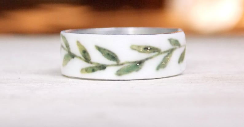 May include: A white ceramic ring with a green leafy design. The leaves have a subtle gold glitter detail.