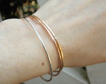 May include: Three gold bangles stacked on a wrist. The bangles are hammered and have a shiny finish. The top bangle is rose gold, the middle bangle is yellow gold, and the bottom bangle is silver.