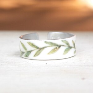 May include: A white metal ring with a delicate green leaf design. The leaves are painted with a watercolor effect and have a subtle gold shimmer.
