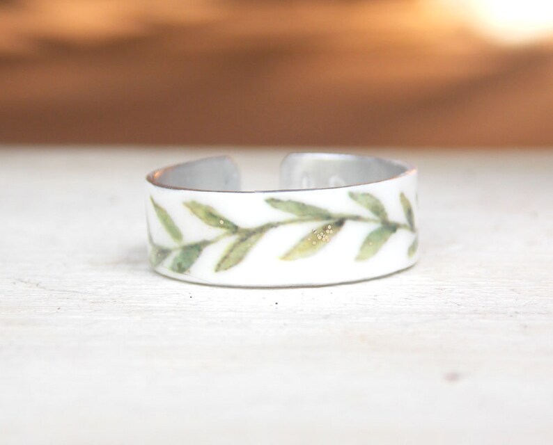 May include: A white metal ring with a delicate green leaf design. The leaves are painted with a watercolor effect and have a subtle gold shimmer.