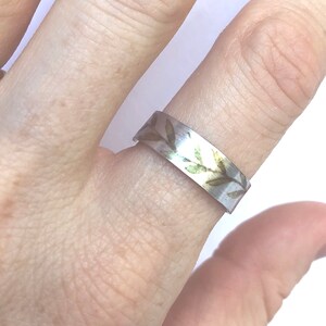 May include: A silver ring with a delicate design of green leaves and gold accents. The ring is worn on a finger.