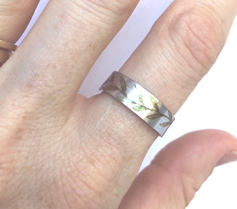 May include: A silver ring with a delicate design of green leaves and gold accents. The ring is worn on a finger.