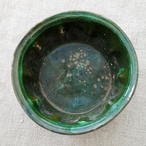 May include: A round, deep bowl with a dark green glazed interior featuring a scalloped rim. The bottom of the bowl displays a mottled, weathered texture with visible brown and dark green spots, showing a glossy finish throughout the surface.