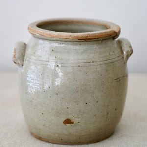 May include: A small, round, off-white ceramic jar with two small handles and a wide rim. The jar has a slightly textured surface with subtle variations in colour and a few small brown spots. The rim and handles have a darker, brownish hue.