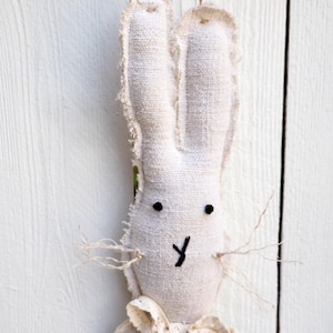 May include: A white fabric bunny rabbit with black eyes and a stitched smile. The bunny is hanging from a white wooden fence with a white lace bow.