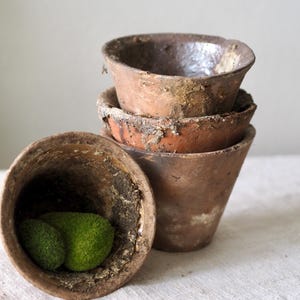May include: A collection of weathered, terracotta-colored clay pots. One pot is on its side, revealing two green moss balls. The other pots are stacked, showing their aged, textured surfaces. The pots have a rustic, vintage aesthetic.