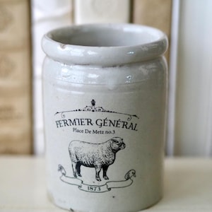 May include: A cylindrical off-white ceramic crock featuring a black printed graphic of a sheep. The text reads FERMIER GÉNÉRAL, Place De Metz no.3, and 1873. The vessel has a smooth, glazed finish with a thick, rounded rim at the top opening.