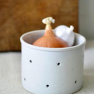 May include: A white ceramic onion and garlic keeper with several small holes. An onion and a garlic bulb are inside the container. The background is a wooden cutting board.