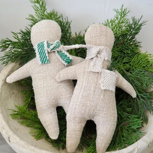 May include: Two beige fabric dolls with simple stitched features and outstretched arms. One doll wears a green and white striped scarf, the other a cream-colored scarf. They rest in a rustic, off-white bowl, surrounded by green sprigs.