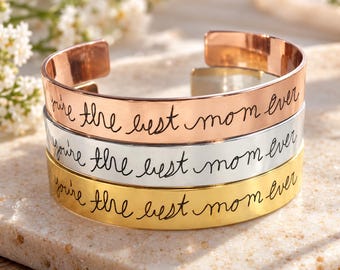 May include: A stack of three metal cuff bracelets in rose gold, silver, and gold tones. Each bracelet features the engraved black cursive text "you're the best mom ever" centered on the front surface. The bracelets have an open back design.