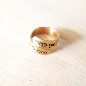 Puede incluir: Un anillo de metal dorado con diseño abierto y acabado liso y pulido. La banda está grabada con texto en hebreo negro que dice 'Ani L'dodi V'dodi Li' en los extremos superpuestos del anillo, creando un diseño envolvente y continuo.