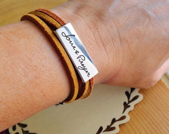 May include: A wrap bracelet featuring two strands of mustard-yellow suede leather cord and a rectangular silver-toned metal bar. The metal bar is engraved with the black cursive text Love & Prayer, positioned horizontally across the leather strands.