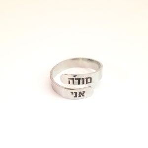 May include: A silver-toned bypass ring featuring an open, adjustable band design. The two ends of the ring are engraved with black Hebrew text, specifically the words "מודה" on the upper terminal and "אני" on the lower terminal, creating a layered look.