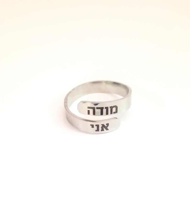 May include: A silver-toned bypass ring featuring an open, adjustable band design. The two ends of the ring are engraved with black Hebrew text, specifically the words "מודה" on the upper terminal and "אני" on the lower terminal, creating a layered look.