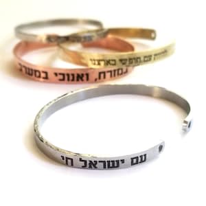Puede incluir: Cuatro pulseras de plata, oro y cobre con texto hebreo grabado en ellas. Las pulseras están apiladas una encima de la otra. El texto en la pulsera inferior dice "Im Shoa'ai Hi".