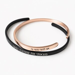 Op de afbeelding: Twee open armbanden, één in matzwart en één in roségoud. De zwarte armband is voorzien van witte Hebreeuwse tekst gegraveerd langs de buitenband, terwijl de roségouden armband zwarte Hebreeuwse tekst gegraveerd heeft op de buitenband.